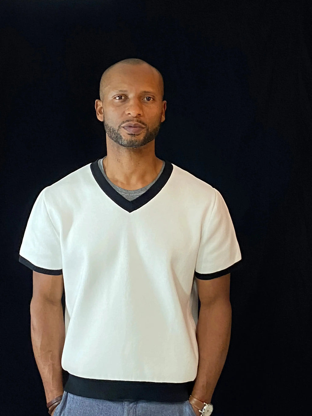 A man wearing a white short-sleeve V-neck t-shirt with black trim on the collar, sleeve cuffs, and hem, paired with gray pants, standing against a solid black background.