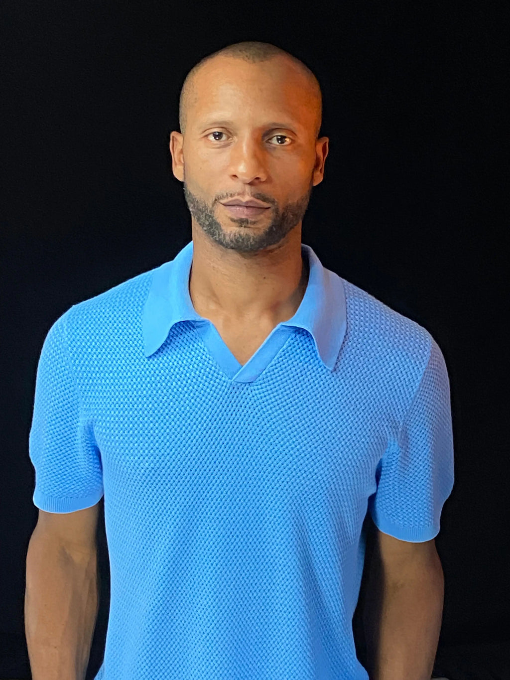 A man wearing a bright sky blue breathable knit short-sleeve polo shirt, standing against a solid black background.