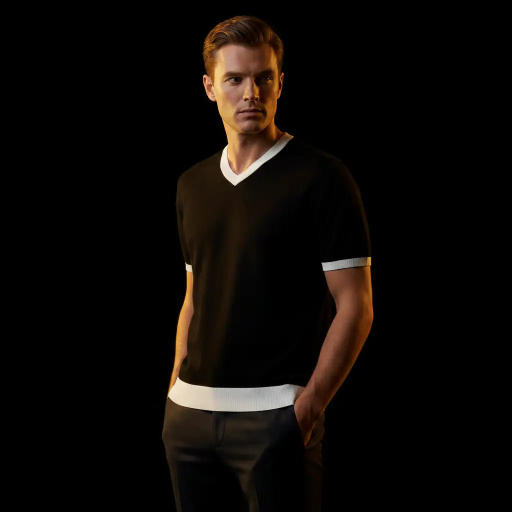 A man wearing a black v-neck t-shirt with white contrast trim at the collar, sleeve cuffs, and hem, paired with dark trousers, posed against a solid black background.